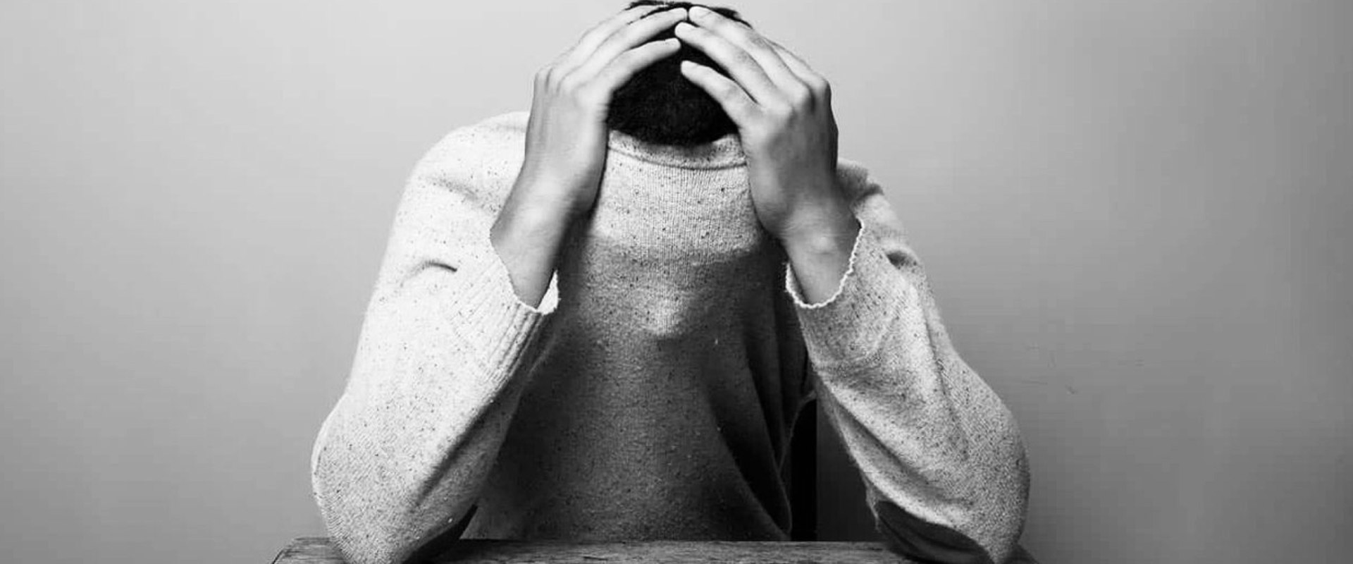 A man sitting on a chair has his face covered by his sweatshirt and holds his hands on his head